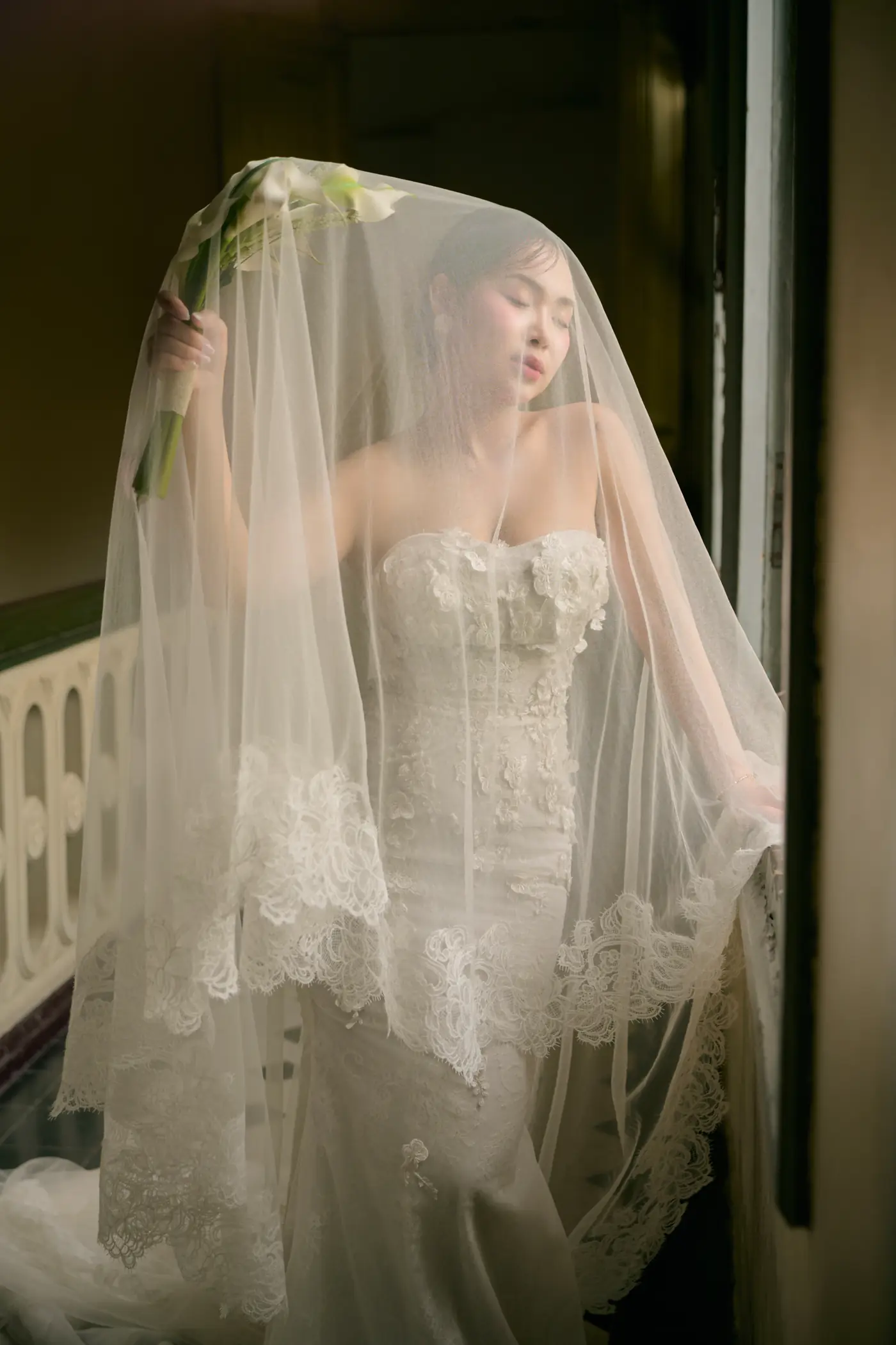 She stands beneath her veil in the window light